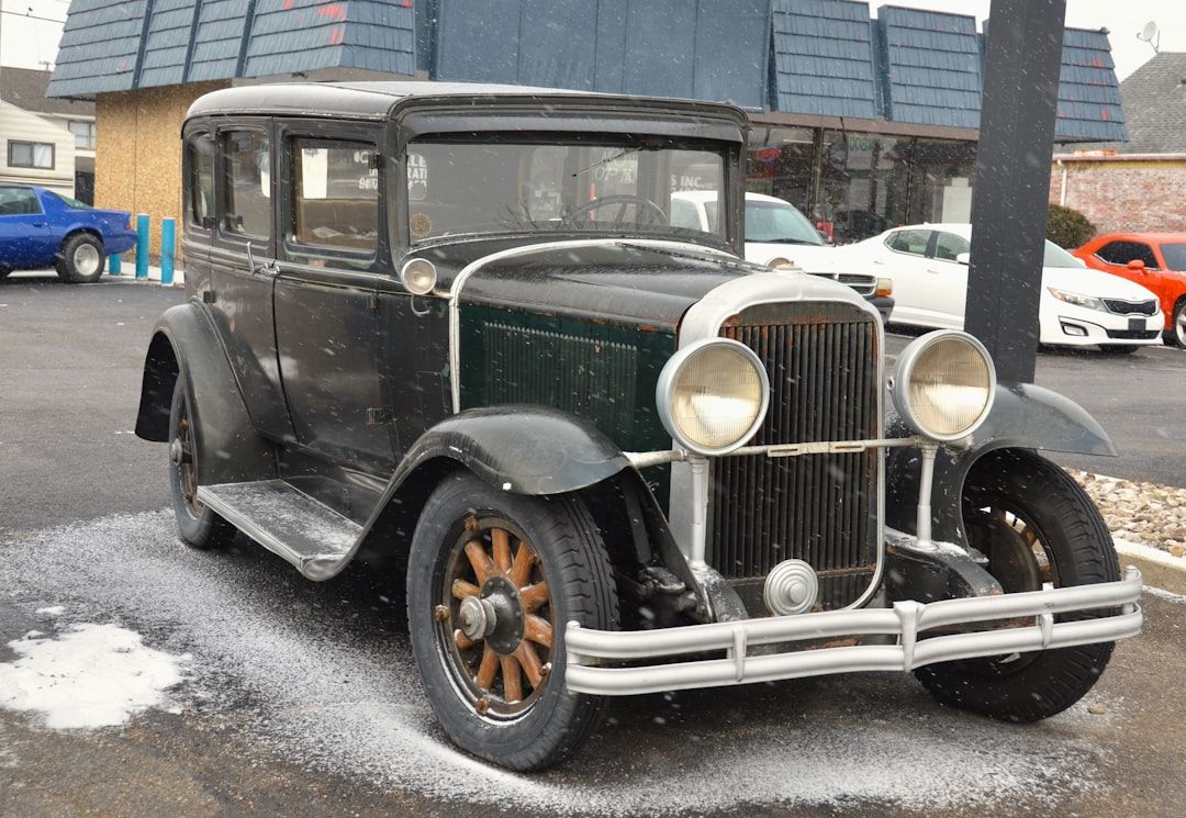 En klassisk veteranbil parkerad på en asfaltplan under lätt snöfall.